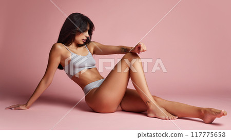 Sitting casually, woman stretches one leg out, her toned body highlighted by gray lingerie, exuding effortless elegance against pink backdrop. Sitting casually, woman stretches one leg out, her toned body highlighted by gray lingerie, exuding effortless elegance against pink backdrop. 117775658