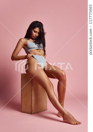 Sitting on wooden block, woman gently pulls on her gray lingerie, revealing her toned midsection and exuding casual confidence against pink background. 117775665