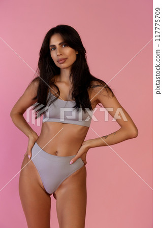 Standing confidently with hands on hips, woman gazes ahead, highlighting her fit physique in gray lingerie against soft pink backdrop. 117775709