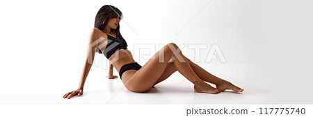 Banner. Woman sits on floor in black lingerie, showcasing her long legs and toned body against white studio background. 117775740