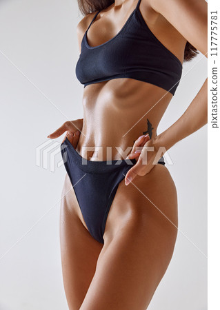 Close-up shot emphasizes woman's wet, toned abs. Model slightly pulling waistband of her black lingerie against white background. Close-up shot emphasizes woman's wet, toned abs. Model slightly pulling waistband of her black lingerie against white background. 117775781