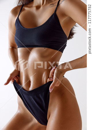 Close-up shot emphasizes woman's wet, toned abs. Model slightly pulling waistband of her black lingerie against white background. 117775782