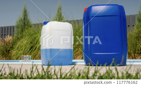 Two large chemical containers are placed poolside with a grass-level view, emphasizing the importance of pool maintenance. Two large chemical containers are placed poolside with a grass-level view, emphasizing the importance of pool maintenance. 117776162