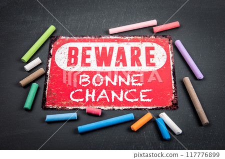 Good Luck. Text in French. Beware sign with text and colored pieces of chalk on a dark chalkboard 117776899