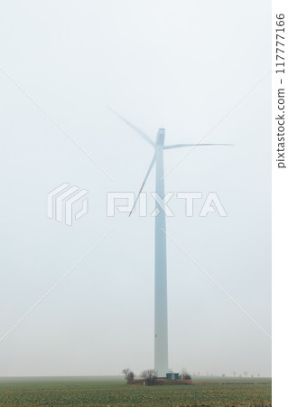 One wind turbine mill in field at foggy misty rainy countryside field low poor visibility on cold spring autumn morning. Seasonal bad rainy weather. Wind mill turbine farm 117777166