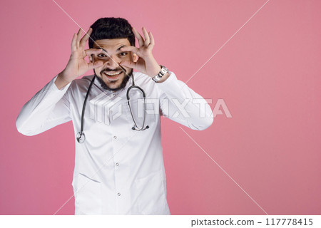 Front view of handsome male doctor wearing medical gown, looking at camera, smiling, touching eyes. Isolated on pink studio background. Front view of handsome male doctor wearing medical gown, looking at camera, smiling, touching eyes. Isolated on pink studio background. 117778415