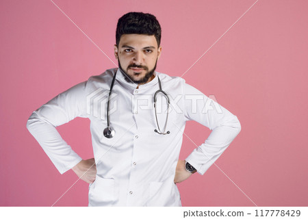 Front view of brunette male doctor, intern standing, holding hands on sides, looking at camera. Isolated on pink studio background. 117778429