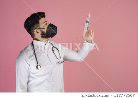 Side view of brunette male doctor with beard standing, looking up, raising hand, holding syringe. Isolated on pink studio background. Side view of brunette male doctor with beard standing, looking up, raising hand, holding syringe. Isolated on pink studio background. 117778507