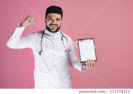 Front view of handsome doctor wearing medical gown, standing, looking at camera, smiling, showing folder. Concept of medicine. Front view of handsome doctor wearing medical gown, standing, looking at camera, smiling, showing folder. Concept of medicine. 117778523