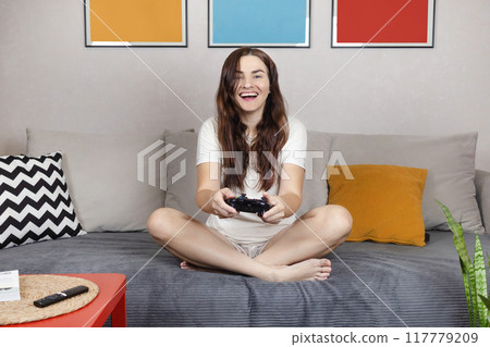 Brunette woman is holding a black jostic, joystick, navigation control. She plays video game on the console. She is smile in home 117779209