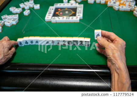A senior holding a Kita tile as he plays mahjong on an electric mahjong table and tries to complete a kokushi musou 117779524