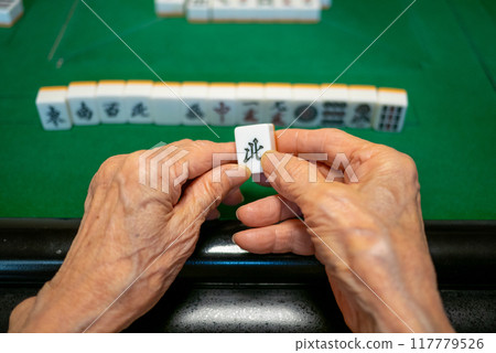 A senior holding a Kita tile as he plays mahjong on an electric mahjong table and tries to complete a kokushi musou 117779526