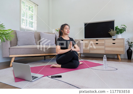 Woman Practicing Yoga at Home on a Pink Mat with Laptop and Water Bottle in Modern Living Room Woman Practicing Yoga at Home on a Pink Mat with Laptop and Water Bottle in Modern Living Room 117783858