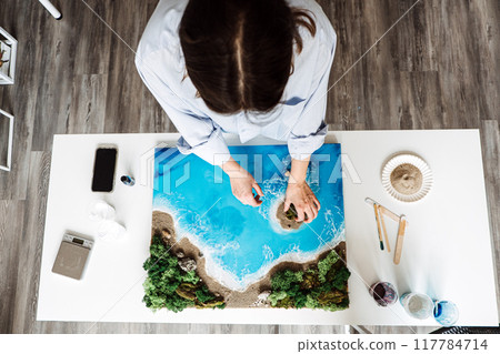 Creating beautiful epoxy resin art at home, featuring a coastal scene with vibrant blue waters, sand, and lush greenery 117784714