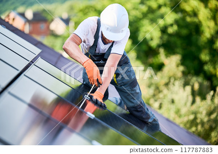 Worker building photovoltaic solar panel system on rooftop of house. Man engineer in helmets and gloves installing solar module with help of hex key outdoors. Renewable energy generation concept. 117787883