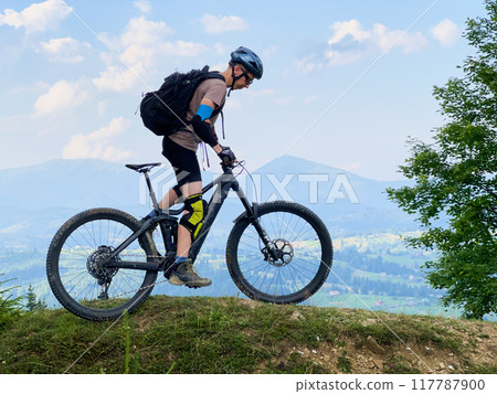 Cyclist man riding electric mountain bike outdoors. Male tourist biking along grassy trail in the mountains, wearing helmet and backpack. Concept of sport, active leisure and nature. 117787900