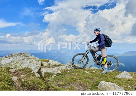 Cyclist man riding electric bike outdoors. Male tourist resting on the top of hill, enjoying beautiful mountain landscape, wearing helmet and backpack. Concept of active leisure. Cyclist man riding electric bike outdoors. Male tourist resting on the top of hill, enjoying beautiful mountain landscape, wearing helmet and backpack. Concept of active leisure. 117787902