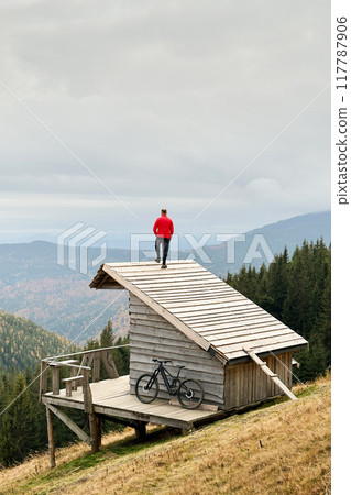 Person resting on edge of steeply sloped wooden rooftop in mountainous area after riding on electric mountain bike on cloudy day. Below the rooftop, there is wooden deck with mountain bicycle. 117787906
