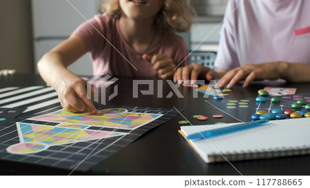 Close-up of young mother and her daughter enjoy a board game at the table indoors, connecting geometric shapes in a tangram puzzle Close-up of young mother and her daughter enjoy a board game at the table indoors, connecting geometric shapes in a tangram puzzle 117788665
