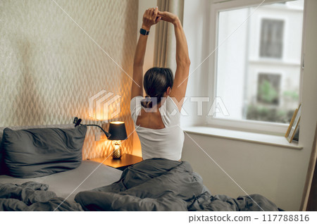 Dark-haired woman feeling relaxed in her bedroom in the morning Dark-haired woman feeling relaxed in her bedroom in the morning 117788816