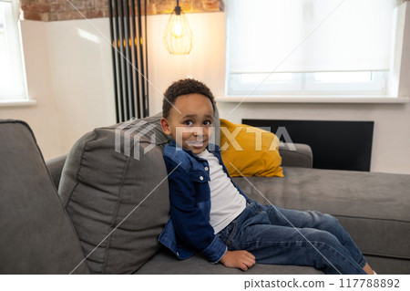 Cute charming black boy sitting on comfortable sofa Cute charming black boy sitting on comfortable sofa 117788892