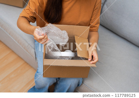 On her sofa, a cheerful lady unboxes a parcel, revealing new headphones, relishing her successful online shopping adventure. 117789125