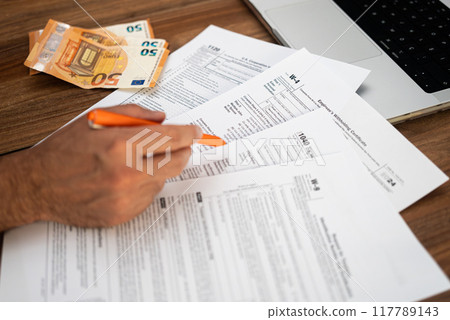 Man entrepreneur engages in paperwork filling complicated tax forms mandatory for business owners sitting at workplace with euro banknotes and laptop in office Man entrepreneur engages in paperwork filling complicated tax forms mandatory for business owners sitting at workplace with euro banknotes and laptop in office 117789143