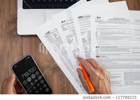Close-up of a man's hands calculating taxes with a smartphone and filling out tax forms on a wooden desk. Financial planning and tax preparation concept.  117789155