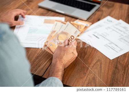 Man handling euro bills while reviewing utility expenses and household finances. Emphasis on financial planning, tax calculations, and managing payments online  117789161