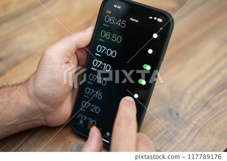 Person adjusting alarms on a smartphone with a wooden background. Digital time management and morning routine planning concept. Suitable for productivity and lifestyle themes  117789176
