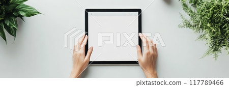 Hands holding a tablet with a blank screen surrounded by green plants on a white surface. Perfect for business presentations and digital content. 117789466