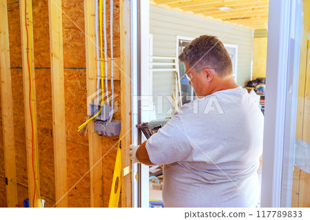 Using pneumatic nail gun, carpenter installs front door in new home 117789833