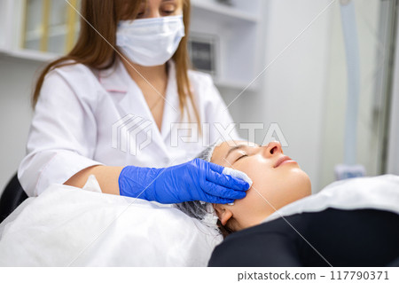 Close-up of a beautician cleaning the skin of beautiful woman with tissues, her hands are in nitrile gloves to maintain the sterility. Perfect cleaning, spa treatment skincare face. 117790371