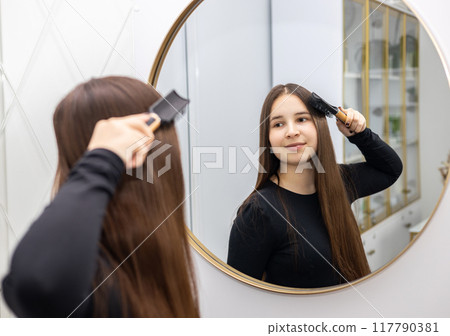 Young woman in a black long sleeve brushes her long hair with a hairbrush while looking in the mirror. 117790381