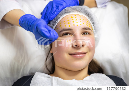 Close-up of a beautiful young woman lying on a treatment table and smiling during a cosmetic procedure. The cosmetologist, wearing sterile gloves, is applying a yellow solution to her face. 117790382