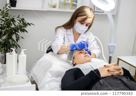 The cosmetologist performs the procedure of dermabrasionon the client's face. The specialist wears a white suit, face mask and medical gloves. Horizontal photo 117790383