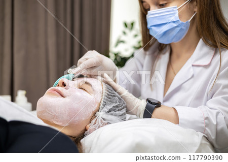 Portrait of a young cosmetologist wearing a surgical mask and white latex gloves, applying a face mask with a spatula on a young woman's face. Portrait of a young cosmetologist wearing a surgical mask and white latex gloves, applying a face mask with a spatula on a young woman's face. 117790390