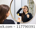 Portrait of a young woman with long hair looking in the mirror with one hand holding a lollipop and with another touching her forehead. The influence of sugar consumption on the skin. 117790391