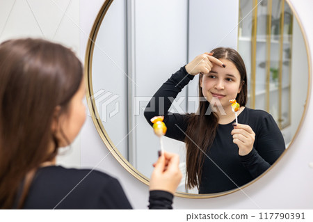 Portrait of a young woman with long hair looking in the mirror with one hand holding a lollipop and with another touching her forehead. The influence of sugar consumption on the skin. 117790391