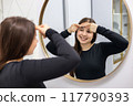 Happy young woman with long hair and a smile on her face looking in the mirror and popping a pimple on her forehead. 117790393