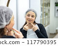 Woman popping acne on chin with hands, dirt or scar on skin. Dermatology, facial wellness and girl in bathroom to squeeze pimple, checking reflection or morning routine 117790395