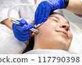 A professional dermatologist wearing medical gloves performs a microdermabrasion procedure on a smiling young woman in a professional beauty salon. 117790398