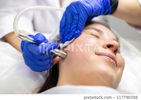 A professional dermatologist wearing medical gloves performs a microdermabrasion procedure on a smiling young woman in a professional beauty salon. A professional dermatologist wearing medical gloves performs a microdermabrasion procedure on a smiling young woman in a professional beauty salon. 117790398