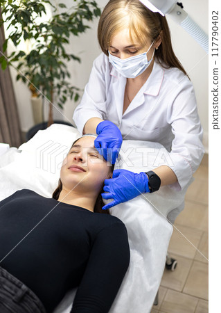Beautician wearing blue nitrile gloves, putting on a hair cap on her client's head to prepare for a cosmetic procedure. 117790402