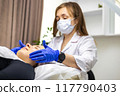 Beautiful young woman enjoying a face massage, lying down on a treatment table in a beauty salon. The beautician is wearing blue nitrile gloves and a face mask. 117790403