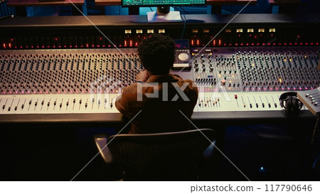 African american music producer finishing his new track in professional studio, recording and editing a song in post production. Skilled audio engineer mixing and mastering tunes. Camera B. 117790646