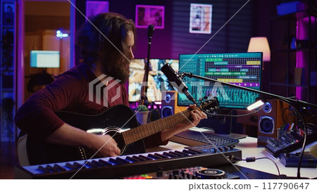 Music producer singing song and playing acoustic guitar, working in his personal home studio to record a new melody. Audio engineer recording tunes, editing with mixing console soundboard. Camera B. 117790647