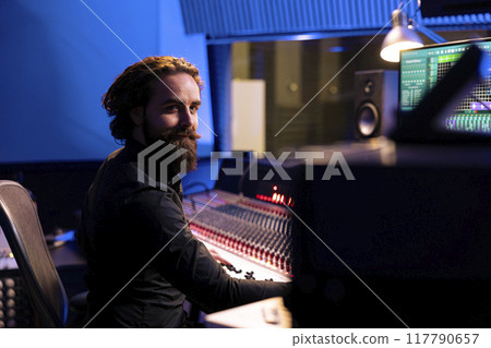 Portrait of sound designer working with mixing console and daw software on pc in professional studio, producing tracks for a pop album. Talented producer editing new music in control room. Portrait of sound designer working with mixing console and daw software on pc in professional studio, producing tracks for a pop album. Talented producer editing new music in control room. 117790657