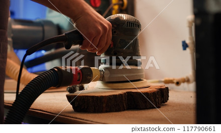 Cabinetmaker at workbench using orbital sander with fine sandpaper on lumber to achieve smooth finish. Woodworking specialist uses angle grinder on wood for professional results, close up, camera A 117790661