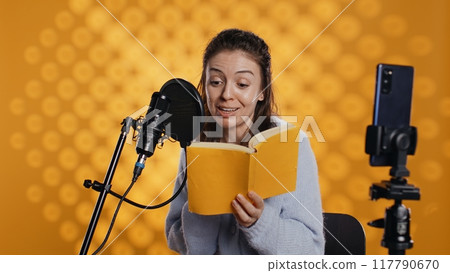 Media star filming vlog at work, reading book to record audiobook, studio background. Voice actor doing content creation while producing digital recording on novel using microphone, camera B 117790670